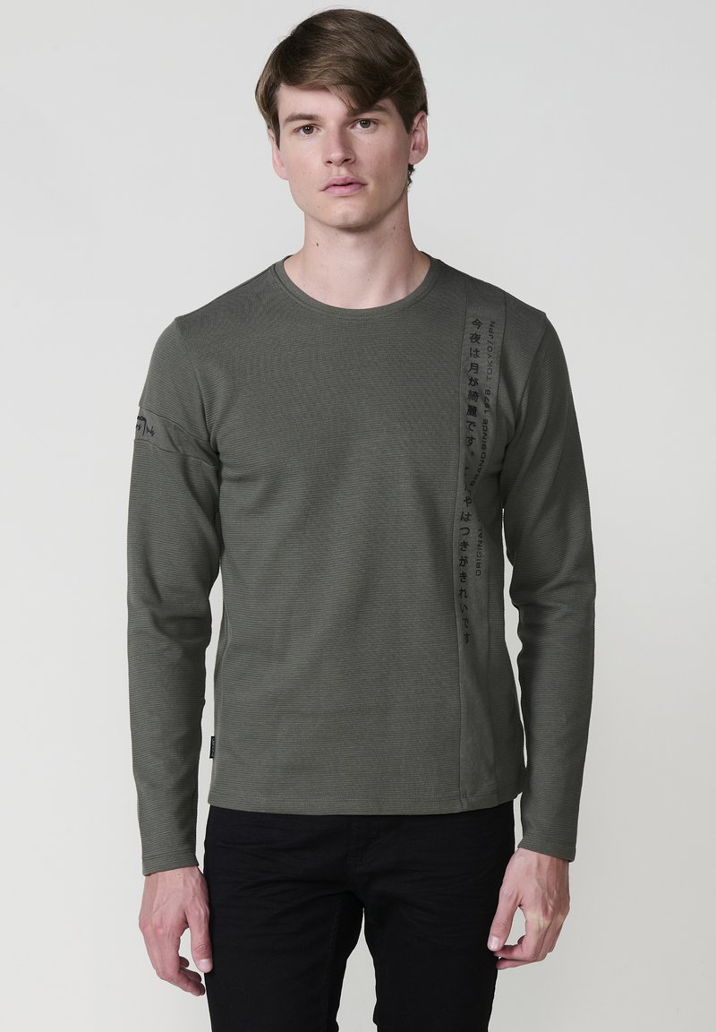 Maslinasto zeleni dugi rukav T-shirt s rebrastom teksturom, okruglim izrezom i vertikalnim crnim natpisom s lijeve strane; kombinirati s crnim hlačama.