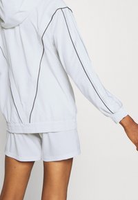 Vit lättviktsjacka med huva, med svart piping längs ärmarna och elastisk midja, stylat med matchande shorts.