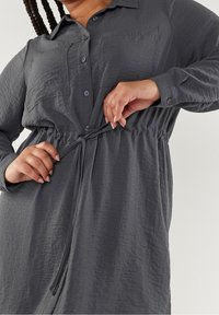 Grijze, knoopsluiting jurk van textuurstof, met twee borstzakken en een verstelbare tailleband voor een getailleerd silhouet. Lange mouwen met knoopmanchetten.