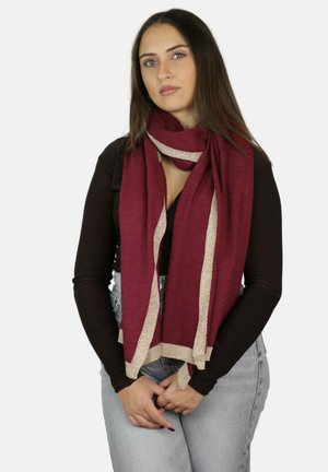 Jonge vrouw met lang bruin haar, gekleed in een donker top met lange mouwen, lichtblauwe spijkerbroek en een bordeauxrode sjaal met beige rand, poserend met in elkaar gewoven handen.