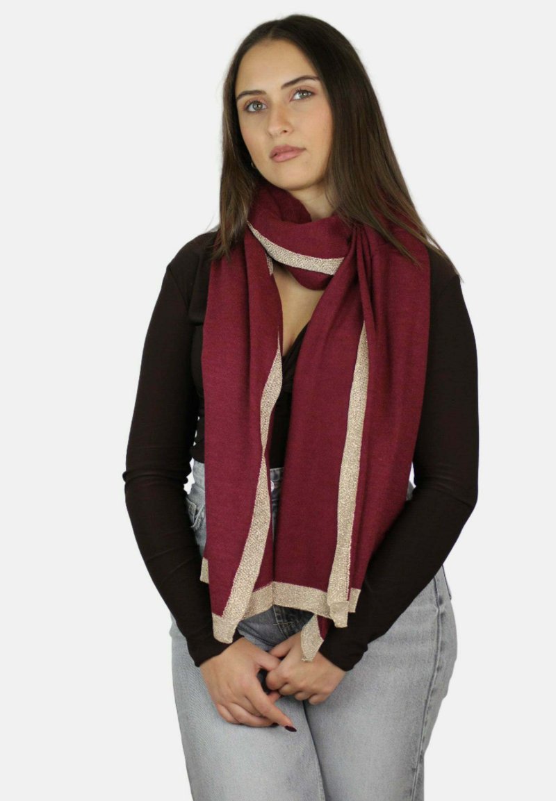 Młoda kobieta z długimi brązowymi włosami, ubrana w ciemną bluzkę z długim rękawem, jasnoniebieskie dżinsy oraz bordowy szalik z beżową lamówką, pozująca z splecionymi rękami.