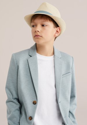 Niño con blazer azul claro y camiseta blanca, mirando de lado, con un sombrero fedora beige con una banda azul sobre un fondo liso.