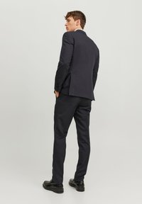 Completo grigio scuro con giacca aderente e pantaloni a gamba stretta. Tessuto liscio, con uno spacco centrale dietro e scarpe eleganti nere lucide.
