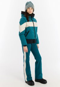 Tyrkysová nafukovacia bunda s krémovými pruhmi, čiernym zipsom a lemom, v kombinácii s tyrkysovými nohavicami s krémovými bočnými pruhmi a čiernou obuvou.