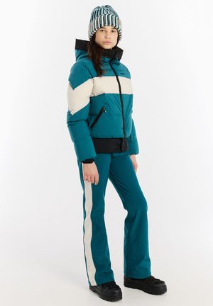 Türkiser Pufferjacke mit cremefarbenen Streifen, einem schwarzen Reißverschluss und Saum, kombiniert mit türkisfarbenen Hosen mit cremefarbenen Seitenstreifen und schwarzem Schuhwerk.