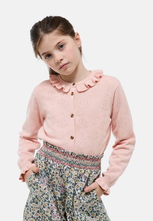 Jeune fille aux cheveux bruns portant un cardigan à col volanté rose et un pantalon taille haute à fleurs, les mains dans les poches, sur fond blanc.