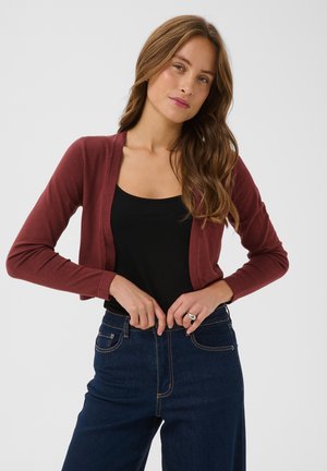 Jeune femme aux longs cheveux ondulés portant un cardigan bordeaux, un top noir et un jean bleu foncé taille haute, ajustant sa taille.