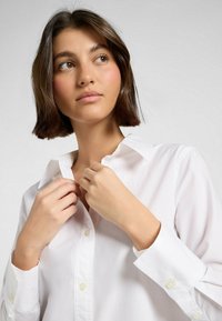 Witte button-up shirt met een klassieke kraag, met een gladde textuur en glanzende witte knopen, gedeeltelijk dichtgeknopt bij de kraag.