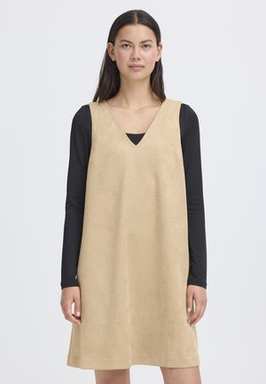 Femme portant une robe beige sans manches par-dessus une chemise noire à manches longues, debout devant un fond clair uni, regardant droit devant avec une expression neutre.