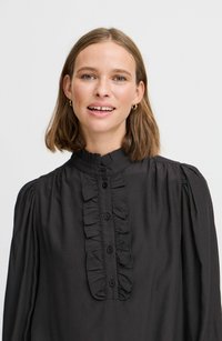 Schwarze Bluse aus leichtem Stoff mit rundem Kragen, Rüschendetail auf der Vorderseite und Knopfverschluss. Puffärmel sorgen für Textur und Form.