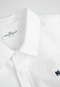 Biała koszula z kołnierzykiem, wykonana z teksturowanej tkaniny, z granatowym haftowanym logo, zapięciem na guziki oraz metką z napisem "Cocodrillo" i szczegółami rozmiaru.