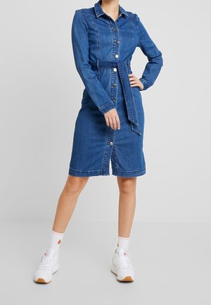Vrouw draagt een knielange spijkerjurk met knopen, lange mouwen en een riem, gecombineerd met witte sokken en witte sneakers.