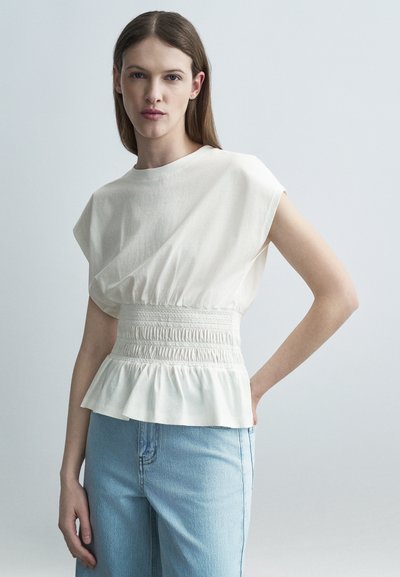 Femme aux cheveux bruns raides portant un haut blanc sans manches à taille froncée et un jean bleu clair, debout devant un fond uni.