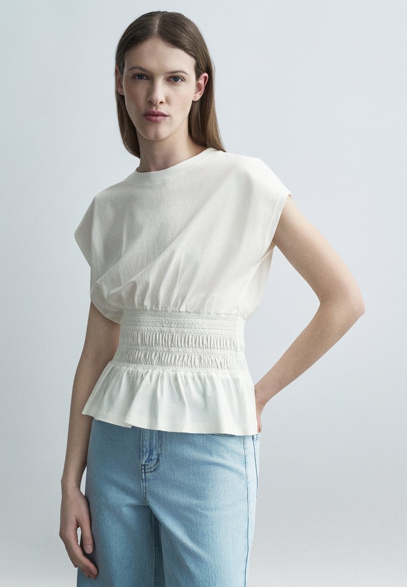 Γυναίκα με ίσια καστανά μαλλιά που φοράει λευκή αμάνικη μπλούζα με σφηκοφωλιά στη μέση και ανοιχτόχρωμο μπλε τζιν, όρθια μπροστά σε ένα απλό φόντο.