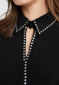 Chemise noire à col avec des clous argentés dessinant le contour du col, du plastron et une ouverture en forme de trou de serrure au niveau de l'encolure, portée par une personne aux cheveux bruns.