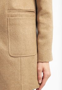 Béžový vlněný kabát s velkou náprsní kapsou zaoblených okrajů, měkkou texturou a čistými liniemi, zachycený z bočního pohledu střední délky.