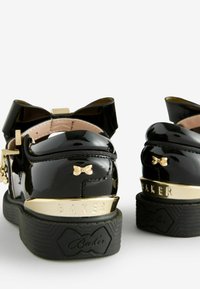 Scarpe in pelle verniciata nera con un accento a fiocco, hardware dorato e una suola in gomma nera texturizzata. "BAKER" impressa sul tallone.