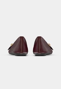 Μπρονζέ μπαλαρίνες από γυαλιστερό δέρμα με στρογγυλεμένη μύτη, επίπεδη σόλα και διακοσμητικό φιόγκο στο μπροστινό μέρος. Λεία υφή και λαμπερό τελείωμα.