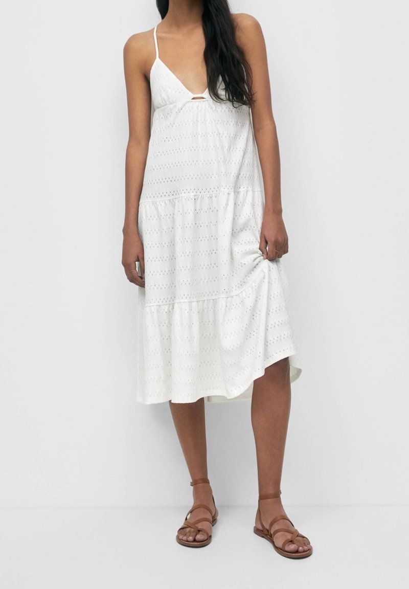 Bílé šaty bez rukávů s výstřihem do V, s předním výřezem a výšivkou na okraji. Vyrobené z texturované tkaniny s očkovými detaily. Hnědé sandály na řemínky.