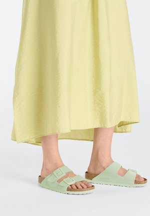 Sandali in suede verde chiaro con due cinturini regolabili, soletta in sughero e suola in gomma testurizzata, abbinati a un abito giallo fluido.