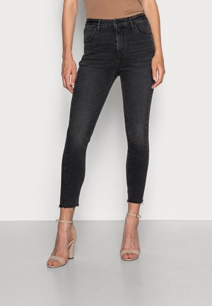 Femme portant un jean skinny noir taille haute et des sandales beiges à bride cheville avec talon bloc, debout devant un fond clair uni.
