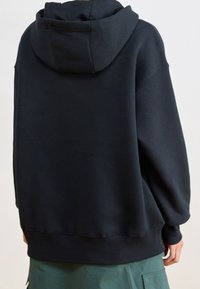 Person som bär en lös svart huvtröja och gröna byxor, sedd bakifrån mot en enkel ljus bakgrund.