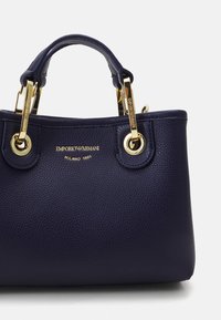 Námořnická kožená kabelka s texturovaným povrchem, zlatými detaily, dvojitými uchy a embosovaným logem na přední straně.