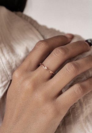 Twisted rose gold ring on a hand, featuring a smooth texture and subtle leaf-like accents, against a soft, neutral fabric background.
