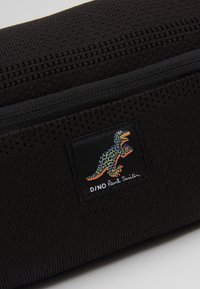 Article en tissu texturé noir avec un patch dinosaure coloré et le texte "D/NO Paul Smith" sur le devant.