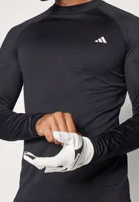 Pitkähihaine musta puristuspaita, jossa on pyöreä kaula-aukko ja valkoinen logo. Mallilla on päällä valkoiset ja mustat käsineet, ja hän ulottaa toista kättään.