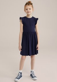 Rochie bleumarin cu mâneci scurte cu volane, corp ajustat și fustă evazată. Asortată cu adidași blue high-top și sosete albe până la gleznă.