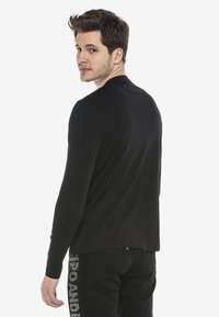 Schwarz langärmliges Shirt mit rundem Ausschnitt, glatter Textur und minimalistischem Design, kombiniert mit schwarzen Jeans mit dekorativer Schriftzug.