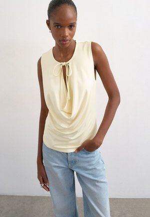 Ung kvinde med mørk hud iført en ærmeløs lysegul top med bindedetalje foran og lyseblå jeans, stående op ad en ensfarvet væg.