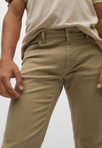 Pantalons en coton beige avec une coupe slim, dotés d'une fermeture à bouton, de passants de ceinture et de poches latérales ; texture lisse et détails de couture minimaux.