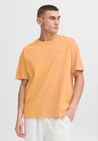 Tričko s krátkým rukávem v jemně oranžové barvě z bavlny s kulatým výstřihem. Obsahuje minimalistické logo v decentním tisku na hrudi. Volný střih.