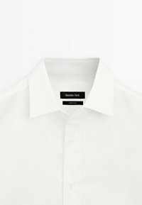 Chemise en coton blanc avec un col classique, une fermeture à boutons, et une étiquette affichant "Massimo Dutti" cousue à l'intérieur. Texture lisse.