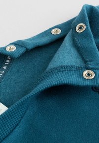 Pull teal avec col boutonné, poignets côtelés et texture douce. Présente des pressions métalliques au niveau de l'encolure pour la fermeture.