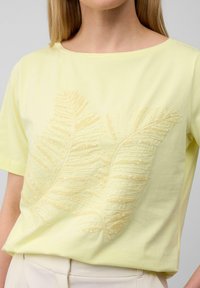 Żółta bluzka z krótkim rękawem z delikatnym haftem w kształcie liści ze złotych cekinów na przodzie. Gładki, lekki materiał w luźnym kroju.