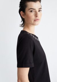 T-shirt nera con scollo rotondo, maniche corte e sottili motivi chiari sulle spalle. Tessuto di cotone morbido, vestibilità rilassata.