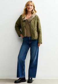 Chaqueta acolchada de color verde oliva con un gran cuello a cuadros, llevada sobre una blusa a cuadros y unos vaqueros de pierna ancha en azul oscuro, combinada con botas negras.