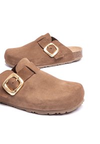 Sabots en daim marron clair avec une large sangle et un accent de boucle en or. Semelle intérieure profilée et semelle en caoutchouc texturé pour une bonne adhérence.