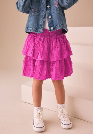 Bambino che indossa una gonna a balze rosa acceso, giacca di jeans con dettagli ricamati, sneakers bianche alte, in piedi vicino a scalini bianchi.
