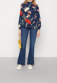 Mörkblå blus med blommönster och långa ärmar, tillsammans med mörkblå utsvängda jeans och vita sneakers, hållandes en gul handväska.