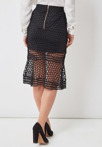 Falda negra con cierre de cremallera y diseño en capas con patrones de crochet intrincados en el dobladillo. Llevada con zapatos de tacón negros.