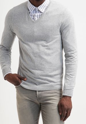 Mann trägt einen hellgrauen V-Ausschnitt-Pullover über einem weiß- und graustreifen Hemd, kombiniert mit hellgrauen Hosen, die eine Hand in der Tasche hat.