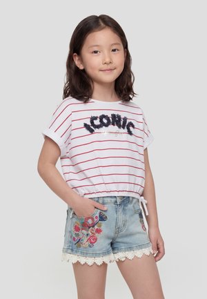 Niña joven con cabello castaño hasta los hombros, vistiendo una camiseta blanca con rayas y texto "ICONIC", y pantalones cortos de mezclilla bordados con ribete de encaje.