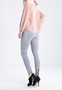 Blouse rose clair à manches longues avec un détail de nœud dans le dos, associée à un jean skinny gris clair avec des chevilles zippées et des chaussures à talons hauts noires.