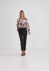 Blusa floral de mangas longas com folhas vermelhas e verdes em fundo branco, combinada com calças pretas de corte ajustado e saltos verdes.