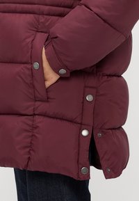Hånd i lommen på en maroon polstret vinterjakke med trykknapdetaljer, båret over mørke denimjeans.