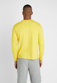 Mann von hinten, der ein gelbes Langarmshirt und graue Shorts trägt, vor einem schlichten weißen Hintergrund.